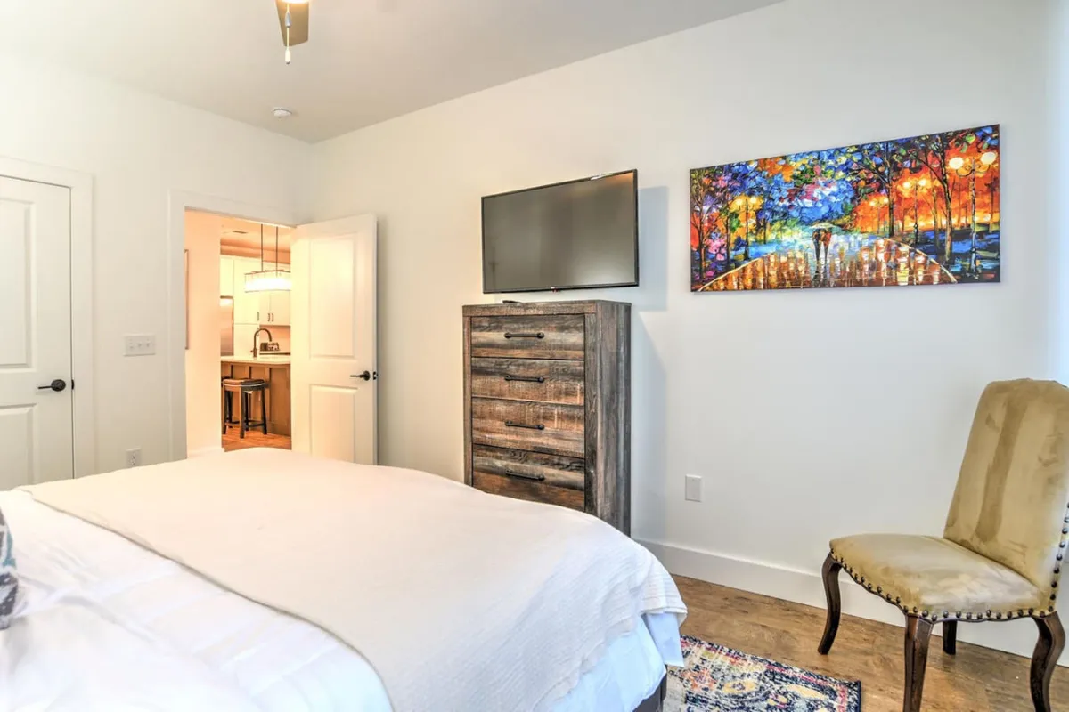 This image presents a serene bedroom featuring a plush bed, a dresser, a TV, wall artwork, and a comfortable chair. The soft lighting and carefully chosen decor create a cozy and inviting ambiance, perfect for relaxation and restful nights.
