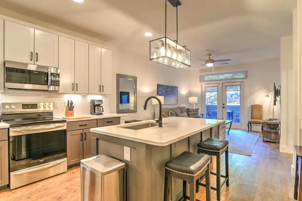 A view of a contemporary kitchen featuring a central island, a modern light fixture, and sleek stainless steel appliances. The kitchen is bathed in natural light, with an open door leading to a patio in the background, suggesting an inviting transition between indoors and outdoors. The space feels both functional and welcoming, ideal for cooking or enjoying a casual meal.