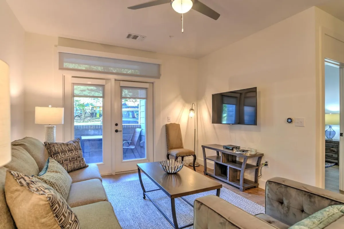 A bright and inviting living room featuring a soft sofa, a practical coffee table, and a television situated opposite the seating area. Sliding doors suggest ample natural light and reinforce the connection between indoor relaxation and the vibrant city outside.